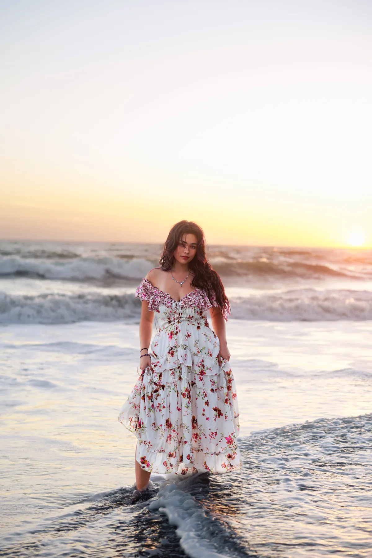 Senior portrait of a young woman in a floral dress standing in the surf at sunset